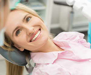 A blond woman with pink shirt smiling up while lying in the dentist's chair